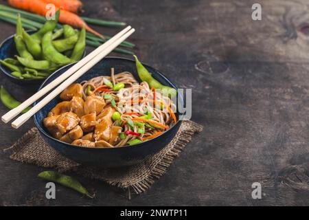 Nouilles asiatiques au poulet, légumes dans un bol, fond rustique en bois. Espace pour le texte. Nouilles soba, poulet teriyaki, édamame, baguettes. Banque D'Images