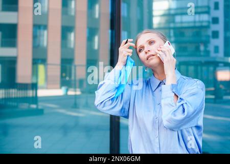 Professionnelle intelligente femme d'affaires caucasienne utilise un téléphone portable en ville à l'extérieur, tenant des documents dans ses mains, vêtue d'une chemise bleue sur le fond d'un centre d'affaires Banque D'Images