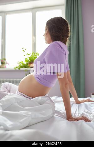 Une femme enceinte s'assoit dans le lit de sa chambre à coucher et regarde par la fenêtre. Femme habillée de pyjama bénéficiant de son état de grossesse. Banque D'Images