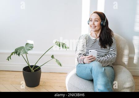 Femme souriante brune utilisant un téléphone portable, écoutez de la musique dans un casque dans le fauteuil à la maison Banque D'Images