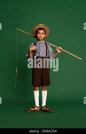 Joli petit garçon, enfant en chemise à carreaux et short suspendu isolé sur fond vert foncé. Concept d'enfance, amitié, rétro Banque D'Images