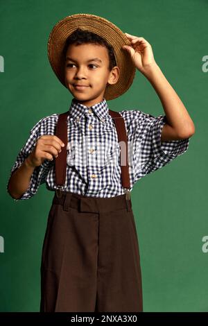 Joli petit garçon, enfant en chemise à carreaux et short suspendu isolé sur fond vert foncé. Concept d'enfance, amitié, rétro Banque D'Images