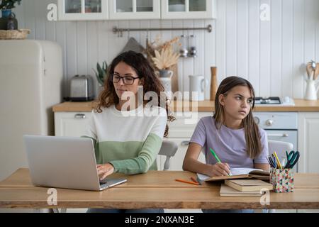 Fille de l'adolescence faisant les devoirs tout en étant assis à la table de cuisine avec le travail de la mère à la maison Banque D'Images