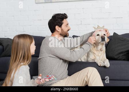 homme barbu souriant portant la couronne de jouet sur le chien près de la fille tenant les ombres à paupières palette, image de stock Banque D'Images