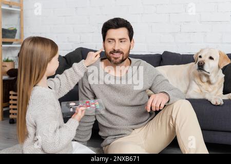 enfant pointant avec le doigt sur le père heureux tout en tenant la palette de maquillage et assis près de labrador chien, image de stock Banque D'Images