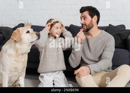 bonne fille portant la couronne de jouet sur la tête tout en étant assis près du labrador et regardant le père appliquant le brillant à lèvres, image de stock Banque D'Images