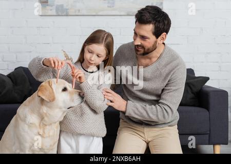 fille heureuse portant la couronne de jouet sur la tête du labrador près du père dans la salle de séjour, image de stock Banque D'Images