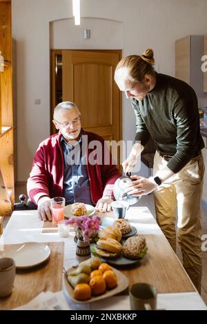 Un jeune homme verse du café pour un père âgé à la retraite avec un handicap pendant le petit-déjeuner à la maison Banque D'Images