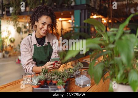 Femme souriante fleuriste portant un tablier tenant le téléphone pendant la journée de travail dans la boutique florale Banque D'Images