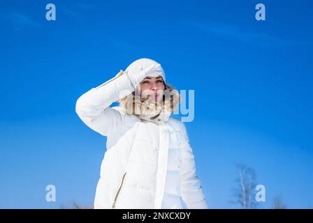 Femme rêveuse en blanc tricoté chapeau et veste tient dans la neige. Main près des yeux. Fond ciel d'hiver Banque D'Images