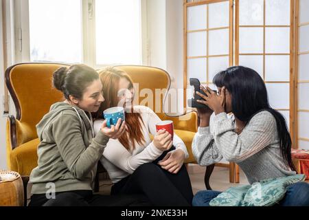 Des amies multiculturelles et gaies qui profitent de leur temps libre à la maison - des jeunes femmes heureuses qui boivent du café ou du thé et qui prennent des photos avec vint Banque D'Images