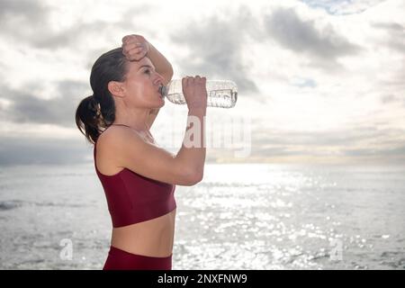 Femme sportive prenant une boisson d'eau d'une bouteille après avoir fait la coloration ou coulu dehors au bord de l'océan Banque D'Images