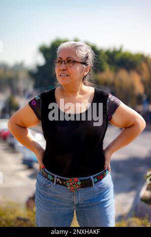 Portrait d'une femme adulte âgée debout avec un paysage urbain flou en arrière-plan, tête tournée sur le côté, vue de face, cheveux gris attachés, vêtements décontractés, bla Banque D'Images