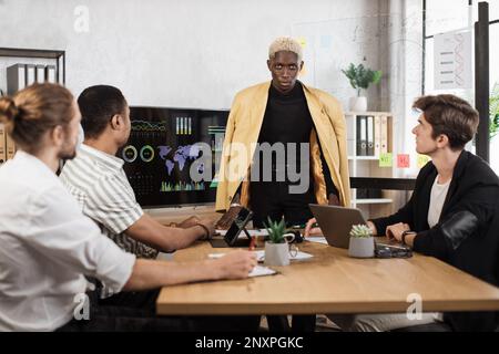 Homme africain se tenant près d'un grand moniteur et d'un tableau de conférence en verre tout en dirigeant une conférence pour des collègues multiraciaux. Les hommes de l'entreprise portent un vêtement formel assis au bureau et à l'écoute du chef d'équipe Banque D'Images