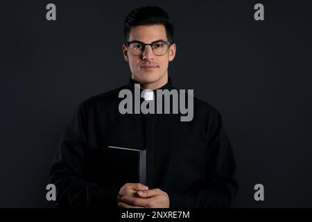 Prêtre en cassock avec Bible sur fond noir Banque D'Images