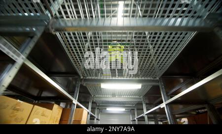 Vue de dessous d'un ouvrier en uniforme lumineux marchant sur le plancher de grille métallique entre les étagères avec des marchandises. Créatif. Équipement d'un entrepôt Banque D'Images