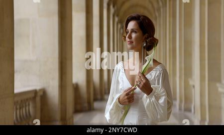 Belle jeune femme romantique dans une longue robe blanche debout près des anciennes colonnes. Action. Belle femme tenant une fleur dans sa main Banque D'Images