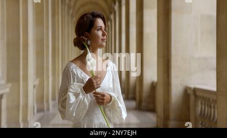 Belle jeune femme romantique dans une longue robe blanche debout près des anciennes colonnes. Action. Belle femme tenant une fleur dans sa main Banque D'Images