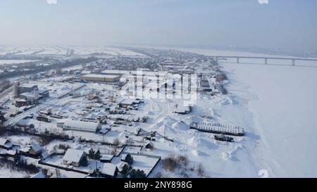Vue aérienne d'une zone industrielle enneigée de la ville. Attache. Vol au-dessus de la ville blanche d'hiver avec la rivière et le pont en arrière-plan Banque D'Images