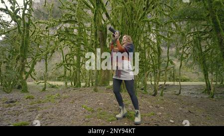 Femme prend des photos sur l'appareil photo dans la forêt. Action. Femme avec appareil photo professionnel dans la forêt verte. Femme prend des photos lors d'une randonnée en forêt avec des arbres mousseux. Banque D'Images