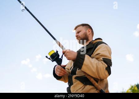 Vue rapprochée à angle bas du pêcheur barbu tenant une tige de coulage portant un imperméable debout sur la rive en attendant des morsures sur la rivière d'eau le jour de l'été. Banque D'Images