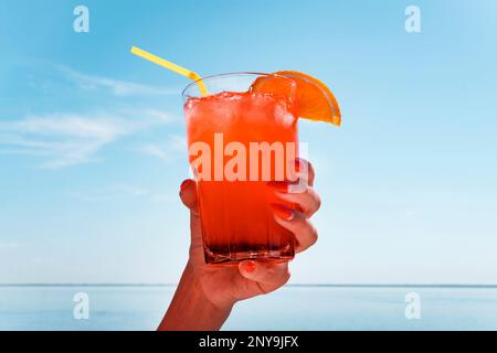 Femme tenant un verre à cocktail contre le ciel bleu et la mer. Une femme boit une boisson rouge. Cocktail de femmes alcoolisées à la main Banque D'Images