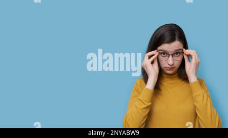 Une jeune fille étudiante souriante qui prend ou met des lunettes sur un fond bleu. La fille voit mieux que d'essayer de nouvelles lunettes prescrites. vérification du soupir Banque D'Images