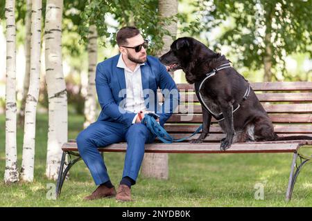 Beau homme en costume d'affaires portant des lunettes de soleil parlant à son chien à la marche Banque D'Images