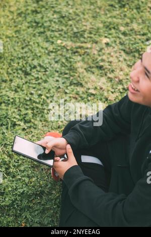 Un adolescent joyeux de descendance latino sourit alors qu'il regarde l'écran de son smartphone. La posture détendue et l'expression gaie du garçon transmettent un sentiment de c Banque D'Images