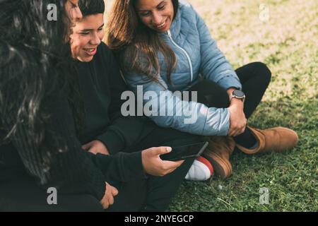 Une famille hispanique passe du temps ensemble dans un parc local lors d'une belle journée ensoleillée. Un adolescent est assis sur l'herbe, tenant son smartphon Banque D'Images