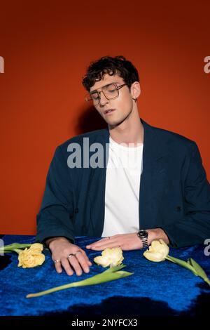 Homme élégant en lunettes assis près des fleurs de tulipe sur fond rouge, image de stock Banque D'Images