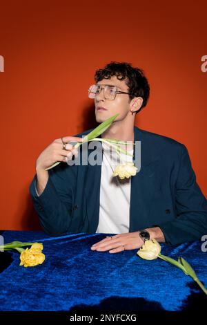 Homme en forme de curly dans un blazer et des lunettes tenant la tulipe sur fond rouge, image de stock Banque D'Images