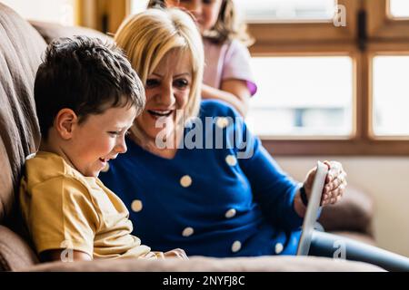 Grand-mère positive et mûre souriant tout en étant assis sur un canapé confortable avec des enfants adorables et un ordinateur portable de surf ensemble Banque D'Images