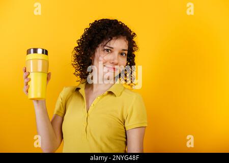 Une jeune femme souriante tient la tasse thermo avec du café ou du thé. Fille en rouge vif et t-shirt jaune. Tasse à café à emporter sans gaspillage chez la femme h Banque D'Images