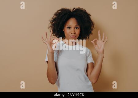 Tout va bien. Jeune femme afro-américaine confiante en t-shirt blanc gardant les deux mains dans un geste convenable, faisant le signe ok tout en se tenant isolé o Banque D'Images