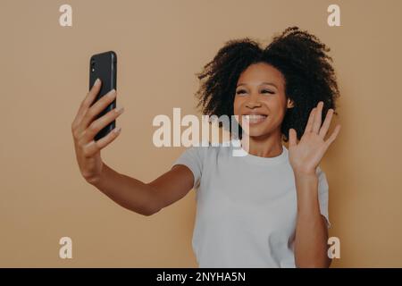 Bonjour ! Belle gaie femme sombre souriant à l'appareil photo sur smartphone moderne, emportant le selfie sur téléphone mobile et se sentant heureuse, en faisant des gestes de salut Banque D'Images