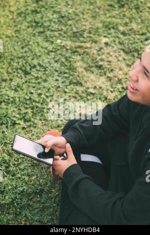 Un adolescent joyeux de descendance latino sourit alors qu'il regarde l'écran de son smartphone. La posture détendue et l'expression gaie du garçon transmettent un sentiment de c Banque D'Images