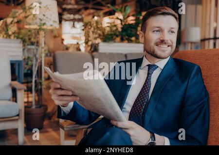 Portrait d'un jeune entrepreneur réussi assis dans des vêtements formels modernes tout en lisant une section de la chronique financière dans le journal, en attente de l'ordre Banque D'Images