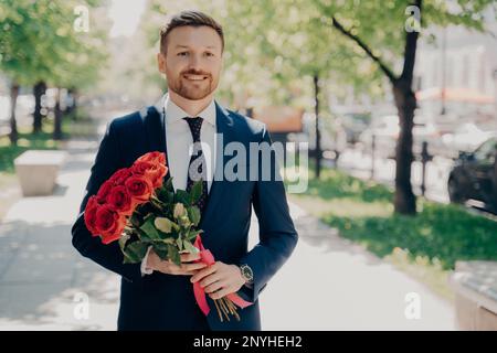 Souriant élégant stubbly gentleman en bleu foncé tuxedo avec grand bouquet de roses rouges marchant le long de l'allée verte dans le parc le jour ensoleillé pour rencontrer son aimé Banque D'Images