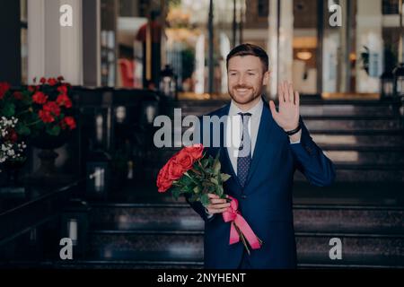 Un bel homme attend dans la rue pour la date dans un élégant costume bleu foncé tenant un bouquet de fleurs roses rouges près du restaurant à l'extérieur tout en levant une main Banque D'Images