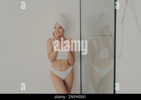 Belle femme debout dans la salle de bains après la douche du matin et l'application de crème hydratante sur le visage, heureuse femme appréciant beauté matin routine à hom Banque D'Images