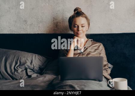 Positive décontracté jeune femme surfant sur Internet pour regarder le film préféré sur ordinateur portable tout en passant une journée de repos à la maison, allongé dans le lit à la maison et looki Banque D'Images