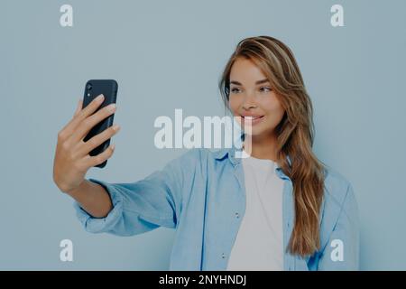 Il est temps de faire un selfie. Magnifique jeune femme dans des vêtements élégants et décontractés avec de longs cheveux teints prenant photo d'elle-même sur un téléphone portable moderne, l'air charmant Banque D'Images