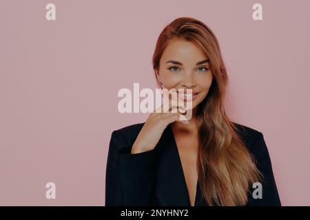 Belle dame de bureau avec de longs cheveux amples et une peau saine. Sourire et toucher légèrement le menton avec les doigts. Porter un élégant blazer noir, un toilettes Banque D'Images