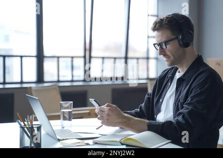 Jeune allemand indépendant dans les écouteurs et les lunettes regardant l'affichage de smartphone dans les mains, en vérifiant les derniers e-mails ou en tapant des SMS de message tout en sittin Banque D'Images