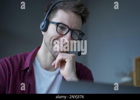 Homme joyeux consultant en centre d'appels parlant allemand dans le micro-casque fonctionne à distance de la maison, souriant poliment tout en communiquant en ligne via ordinateur portable Banque D'Images