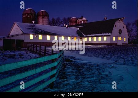 La grange laitière du Connecticut et les silos au crépuscule en hiver avec une lueur chaude à travers les fenêtres contrastant avec le ciel bleu du soir Banque D'Images