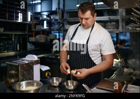 Chef mature en tablier craquant des œufs frais tout en cuisant des nouilles Banque D'Images