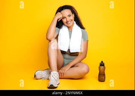 Photo en longueur d'une femme brésilienne ou hispanique, dans des vêtements de sport, entraîneur sportif, avec une serviette sur ses épaules, assis sur un fond jaune isolé, regarde l'appareil photo, souriant amical Banque D'Images