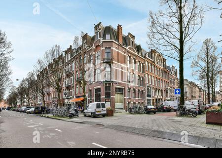 Amsterdam, pays-Bas - 10 avril 2021 : rue de la ville avec beaucoup de voitures garées et de vélos sur le trottoir devant un vieux bâtiment en briques, entouré d'arbres sans feuilles Banque D'Images